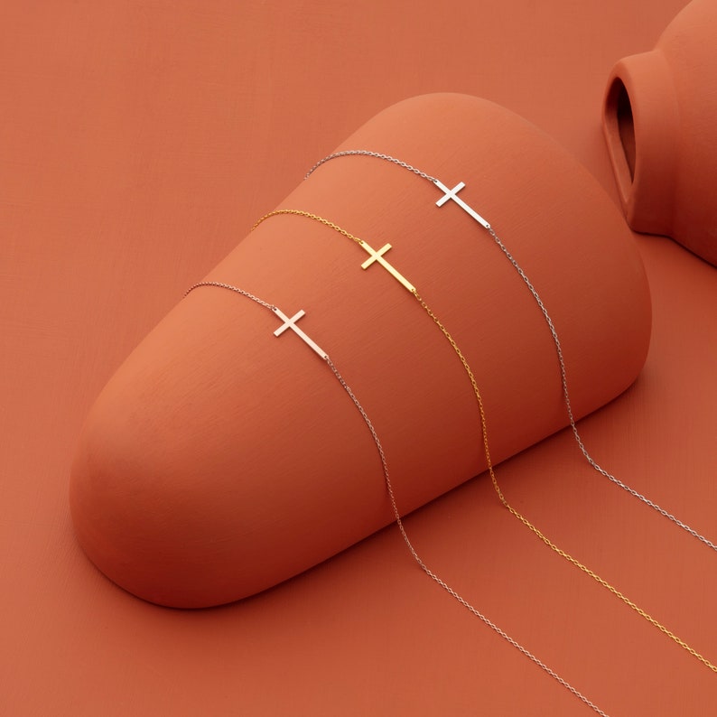 May include: Four delicate necklaces with a simple cross pendant. The necklaces are silver and gold and are displayed on a terracotta background.