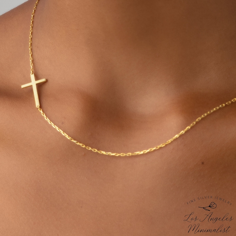 May include: A gold chain necklace with a sideways cross pendant. The necklace is worn on a person's neck. The text "Fine Silver Jewellery by Los Angeles Minimalist" is at the bottom of the image.