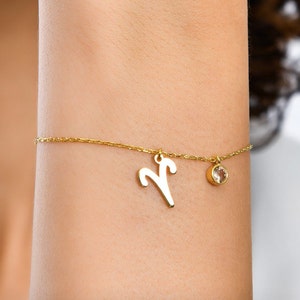 May include: A gold chain bracelet with a small, clear gemstone charm and a larger charm shaped like the astrological symbol for Aries.