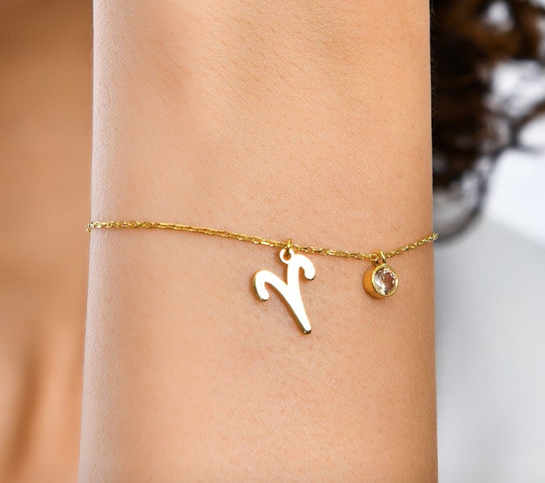 May include: A gold chain bracelet with a small, clear gemstone charm and a larger charm shaped like the astrological symbol for Aries.