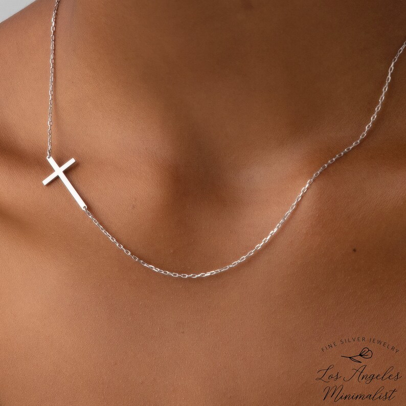 May include: A silver chain necklace with a sideways cross pendant. The necklace is on a person's chest. The text "Fine Silver Jewelry Los Angeles Minimalist" is on the bottom of the image.