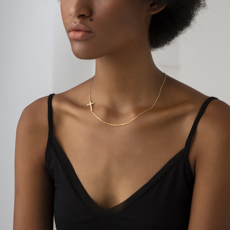 May include: A gold chain necklace with a small, delicate cross pendant. The necklace is worn by a person with dark skin and short, curly hair.