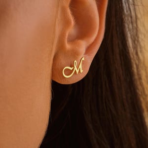 May include: A pair of gold-coloured earrings featuring a cursive letter 'M' design.