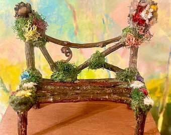 May include: Miniature wooden bench with a rustic design. The bench is adorned with moss, dried flowers, and small pinecones. The bench is brown with a reddish-brown seat and is designed for a fairy garden or similar display.