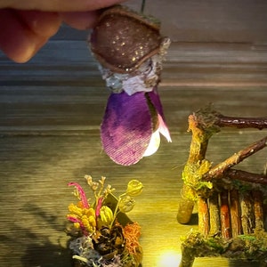 May include: A miniature hanging light fixture with a brown textured cap and purple fabric petals, emitting a warm glow. Below, a small arrangement of dried flowers and moss sits next to a tiny rustic wooden bench made of twigs and moss.