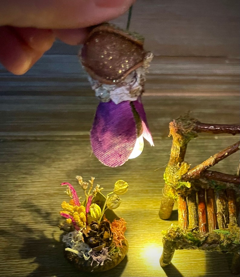 May include: A miniature hanging light fixture with a brown textured cap and purple fabric petals, emitting a warm glow. Below, a small arrangement of dried flowers and moss sits next to a tiny rustic wooden bench made of twigs and moss.