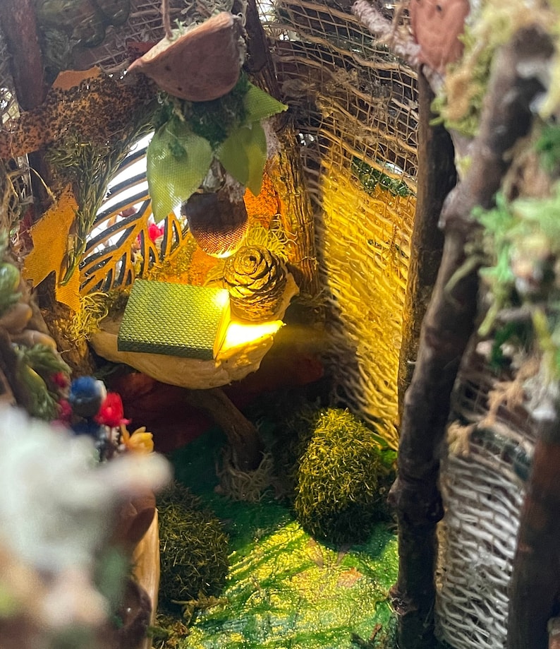May include: A miniature diorama featuring a small green book and a pinecone resting on a shelf. The scene is illuminated by a warm yellow light, surrounded by natural elements including twigs, moss, dried leaves, and a hanging acorn-shaped ornament.