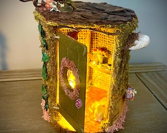 May include: A miniature fairy house with an open door, illuminated from within. The house is made to look like a tree trunk, with a textured bark roof, moss, and decorative flowers. The door has a wreath and a small light.
