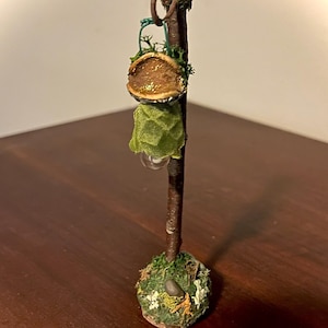 May include: A miniature lamp post featuring a dark brown twig stem, a green fabric shade, and a base decorated with green moss, small white flowers, and a tiny gray stone. The text ForestFindsNFairies is printed at the bottom of the image.