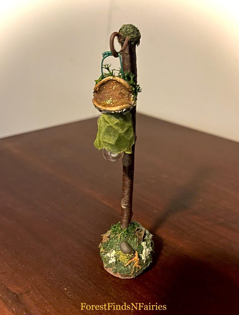 May include: A miniature lamp post featuring a dark brown twig stem, a green fabric shade, and a base decorated with green moss, small white flowers, and a tiny gray stone. The text ForestFindsNFairies is printed at the bottom of the image.