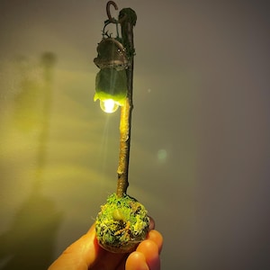 May include: A miniature lamp post featuring a brown twig-like pole anchored in a moss-covered base. A small, green, leaf-shaped shade hangs from a hook at the top, emitting a warm, glowing light from a tiny bulb positioned beneath the foliage.