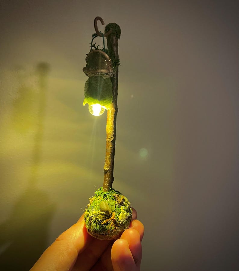 May include: A miniature lamp post featuring a brown twig-like pole anchored in a moss-covered base. A small, green, leaf-shaped shade hangs from a hook at the top, emitting a warm, glowing light from a tiny bulb positioned beneath the foliage.