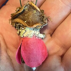 May include: A small decorative ornament featuring a brown, textured cap with gold glitter, a light-colored lichen-like midsection, and a pink fabric petal base. A thin green wire loop is attached to the top for hanging this miniature object.