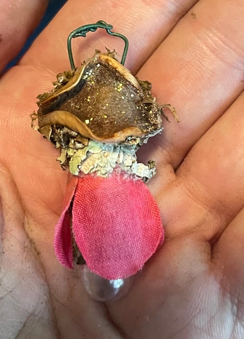 May include: A small decorative ornament featuring a brown, textured cap with gold glitter, a light-colored lichen-like midsection, and a pink fabric petal base. A thin green wire loop is attached to the top for hanging this miniature object.