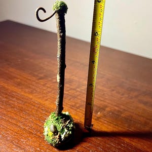 May include: A brown twig stand with a moss-covered base and a metal hook at the top. A yellow measuring tape placed next to the stand indicates a total height of approximately 6.5 inches from the base to the top of the hook.