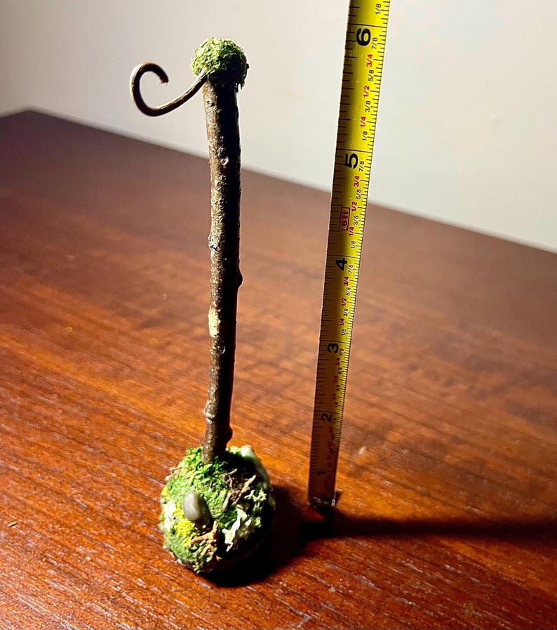 May include: A brown twig stand with a moss-covered base and a metal hook at the top. A yellow measuring tape placed next to the stand indicates a total height of approximately 6.5 inches from the base to the top of the hook.