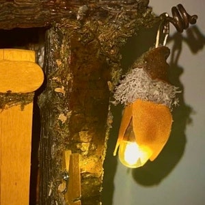 May include: A decorative structure made of dark brown tree bark and green moss featuring a hanging acorn-shaped light fixture. The light glows with a warm yellow hue and is accented by a textured orange fabric cap and light gray lichen-like material.