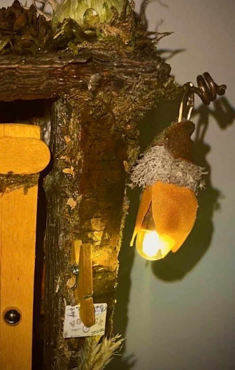 May include: A decorative structure made of dark brown tree bark and green moss featuring a hanging acorn-shaped light fixture. The light glows with a warm yellow hue and is accented by a textured orange fabric cap and light gray lichen-like material.