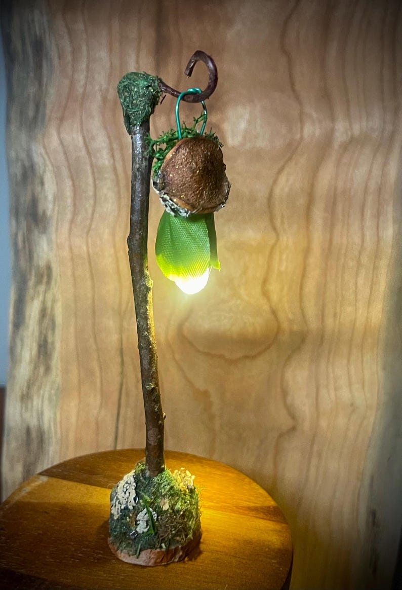May include: A miniature lamp featuring a brown twig-like post standing on a mossy base. A small, brown, textured mushroom cap hangs from a curved hook at the top, with a green fabric leaf element and a glowing white light emitting from below.