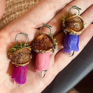 May include: Three small ornaments featuring brown, glitter-dusted acorn caps topped with green moss and a wire loop. Each ornament has a fabric petal base in shades of magenta, light pink, and purple, attached to a small, clear glass bulb at the bottom.