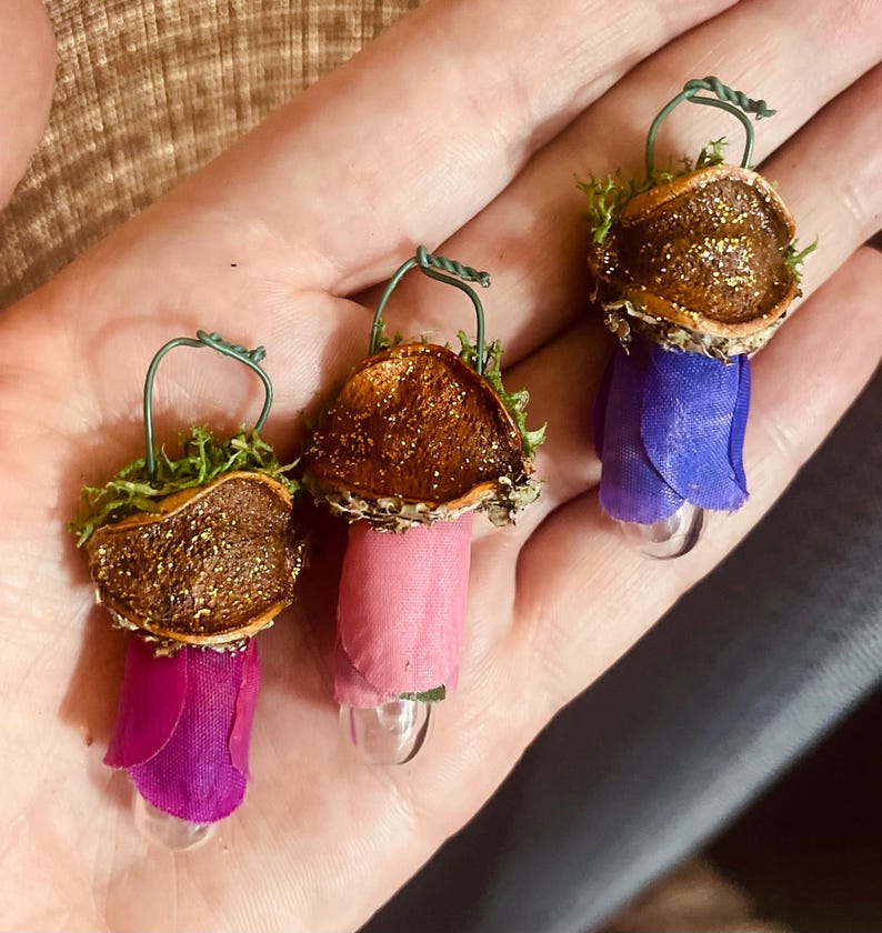 May include: Three small ornaments featuring brown, glitter-dusted acorn caps topped with green moss and a wire loop. Each ornament has a fabric petal base in shades of magenta, light pink, and purple, attached to a small, clear glass bulb at the bottom.