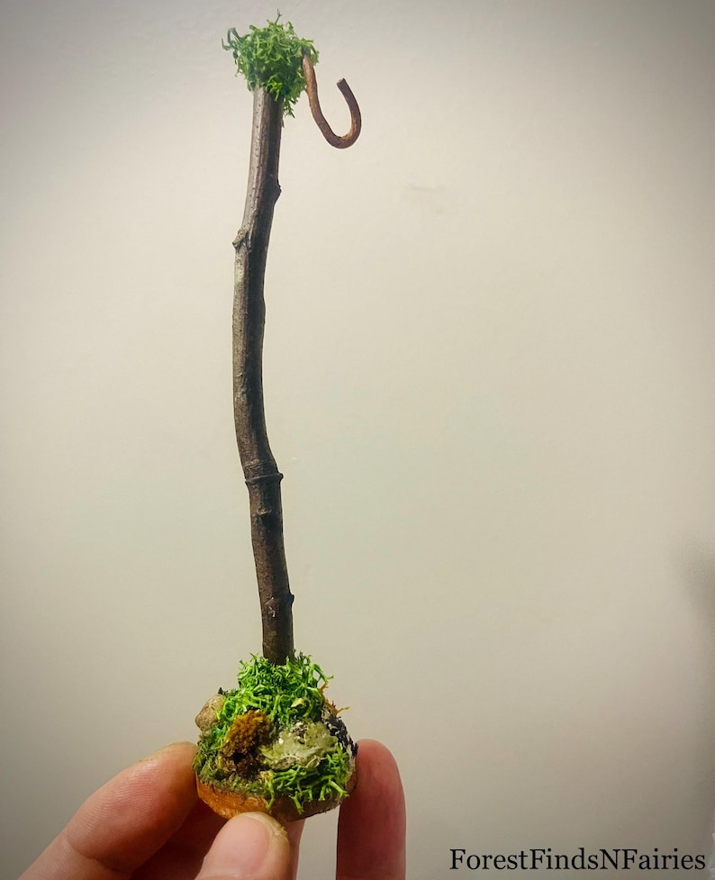 May include: A miniature brown wooden staff standing upright on a circular base covered in green moss and small stones. The top of the staff features a small cluster of green moss and a curved brown hook. The text ForestFindsNFairies is visible.
