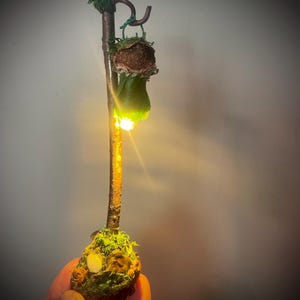 May include: A vertical brown twig stand with a moss-covered base and a curved top hook. A small, textured, bell-shaped object hangs from the hook, emitting a bright, warm yellow light from its base, illuminating the twig and the green mossy ground.