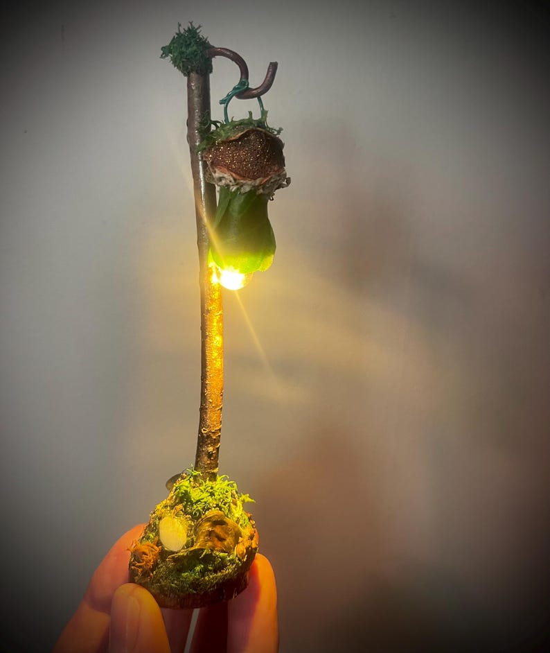 May include: A vertical brown twig stand with a moss-covered base and a curved top hook. A small, textured, bell-shaped object hangs from the hook, emitting a bright, warm yellow light from its base, illuminating the twig and the green mossy ground.