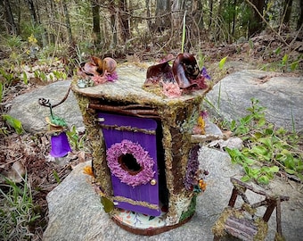 May include: A whimsical fairy house with a purple door and a wreath, set in a natural forest environment. The house is adorned with moss, flowers, and a small wooden chair. The text "ForestFindsNFairies" is visible at the bottom.