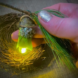 Könnte beinhalten: Ein kleines, grünes, leuchtendes Ornament in Form eines Vogelnests mit einem leuchtenden Licht im Inneren. Das Ornament wird in einer Hand mit silbernem Glitzer-Nagellack gehalten.
