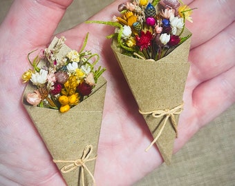 May include: Two miniature bouquets of dried flowers wrapped in brown kraft paper cones. Each cone is secured with a thin, light brown twine tied in a small bow. The floral arrangements feature a mix of yellow, white, pink, red, and blue blossoms.