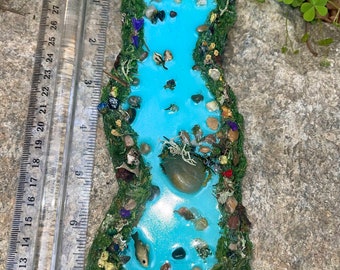 May include: A miniature fairy garden pond with a blue resin water feature, surrounded by green moss, small rocks, and a butterfly figurine.