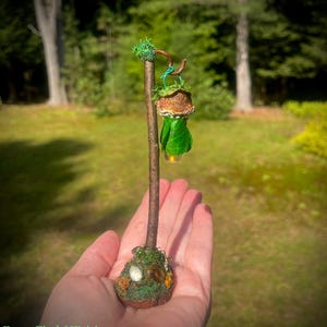 May include: A miniature lamp post crafted from a brown twig, featuring a hanging lantern with a textured brown cap and green leaf-like base. The base is decorated with green moss and small stones. The text ForestFindsNFairies is visible.