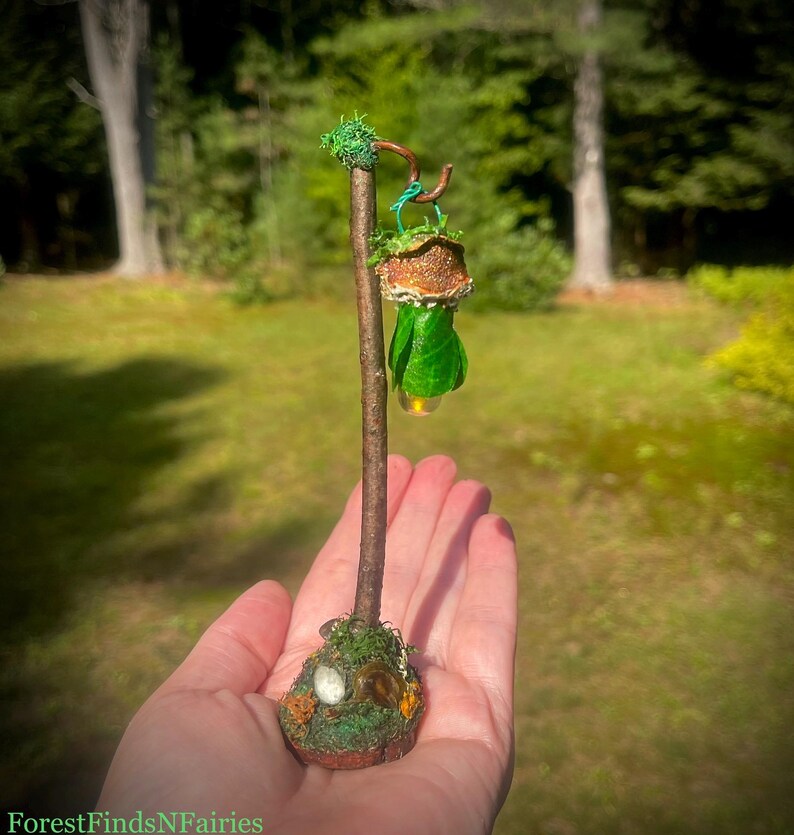 May include: A miniature lamp post crafted from a brown twig, featuring a hanging lantern with a textured brown cap and green leaf-like base. The base is decorated with green moss and small stones. The text ForestFindsNFairies is visible.