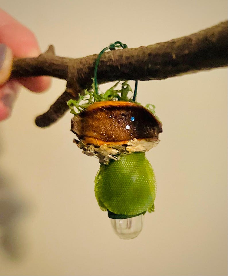 May include: A small hanging ornament featuring a brown acorn cap topped with green moss, suspended by a thin green wire. Below the cap is a bright green fabric sphere with a small, clear, translucent bulb-shaped piece attached at the bottom.