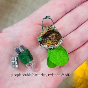 May include: A miniature decorative ornament featuring a dark brown, glittery, acorn-like cap adorned with green moss and two bright green leaves, accompanied by a small clear capsule with a green band, three silver batteries, and text: 3 replaceable batteries, twist on & off.