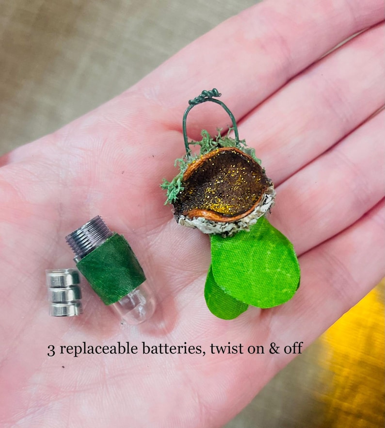 May include: A miniature decorative ornament featuring a dark brown, glittery, acorn-like cap adorned with green moss and two bright green leaves, accompanied by a small clear capsule with a green band, three silver batteries, and text: 3 replaceable batteries, twist on & off.