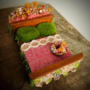 Könnte beinhalten: Ein Miniaturbett mit einem rosafarbenen Juteschlafbezug, Moos-Kissen und einem Blumenkranz am Fußende. Das Kopfteil ist mit getrockneten Blumen verziert.