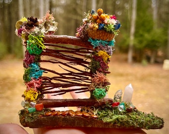May include: A miniature fairy garden fence made of twigs and moss, decorated with colorful beads and flowers. The fence is topped with a small basket filled with dried flowers and berries.