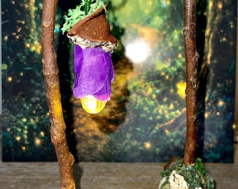 May include: Two miniature lamp posts made of brown twigs with mossy bases. One lamp post features a hanging purple fabric lantern with a glowing yellow light, while the other is a bare post with a mossy top, both designed for miniature displays.