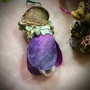 May include: A small decorative ornament featuring a brown textured cap with gold glitter, a light green lichen-covered stem, and purple fabric petals surrounding a small silver metallic sphere at the base, with a thin green wire loop at the top.