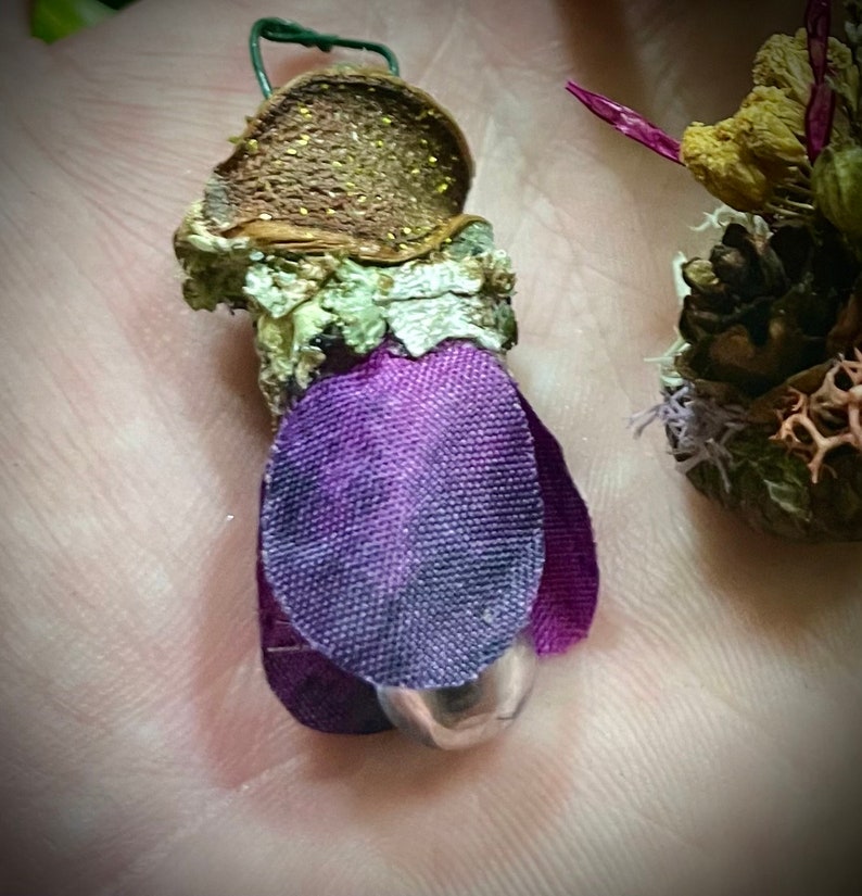 May include: A small decorative ornament featuring a brown textured cap with gold glitter, a light green lichen-covered stem, and purple fabric petals surrounding a small silver metallic sphere at the base, with a thin green wire loop at the top.