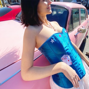 Op de afbeelding: Een vrouw draagt een blauwe satijnen corset met witte sterren en een speelkaartenontwerp. De corset is strapless en heeft een sweetheart-halslijn. De vrouw leunt tegen een roze auto.