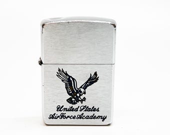 May include: A brushed silver metal flip-top lighter featuring a black graphic of an eagle in flight above the text United States Air Force Academy. The rectangular lighter has a hinged lid and a metallic finish with visible horizontal graining.