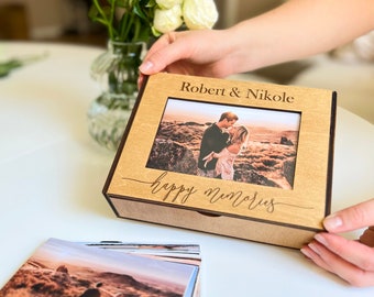 May include: A wooden photo box with a personalized inscription "Robert & Nikole" and the words "happy memories" engraved on the front. The box contains a stack of photos of a couple.