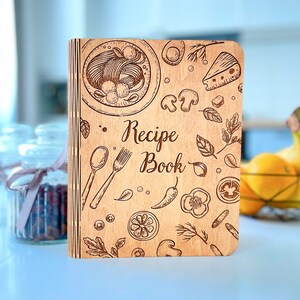 Puede incluir: Un libro de recetas de madera con una cubierta marrón y un diseño de varios alimentos, incluyendo pasta, queso, champiñones y pimientos. El texto "Recipe Book" está grabado en la cubierta.