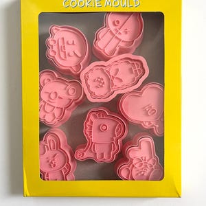 May include: A yellow box with a clear window showing pink plastic cookie cutters shaped like cartoon characters. The box says "COOKIE MOULD" on the top.