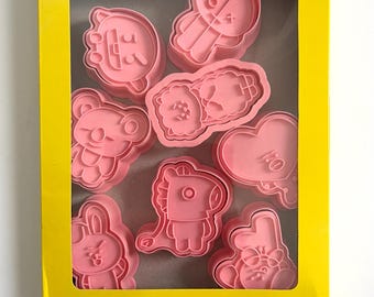 May include: A yellow box with a clear window showing pink plastic cookie cutters shaped like cartoon characters. The box says "COOKIE MOULD" on the top.