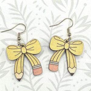 Pode incluir: Brincos de madeira com design de laço e lápis amarelo. Os brincos têm ganchos prateados. A imagem inclui o texto "Glowforge Ready" e "SVG FILE". Produto digital. O texto "not a physical product" também está incluído.