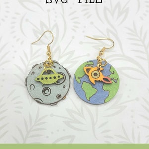 May include: A pair of earrings featuring a moon and a planet with a spaceship on each. The moon is grey with a green spaceship, and the planet is blue with a green and orange spaceship. The earrings are made of wood and have gold hooks.